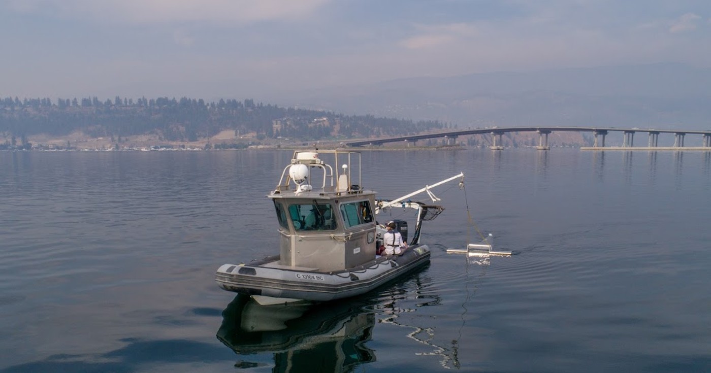 A research vessel skims a manta trawl through the waters of Okanagan Lake with the William R. Bennett bridge in the background on a smoky July day.